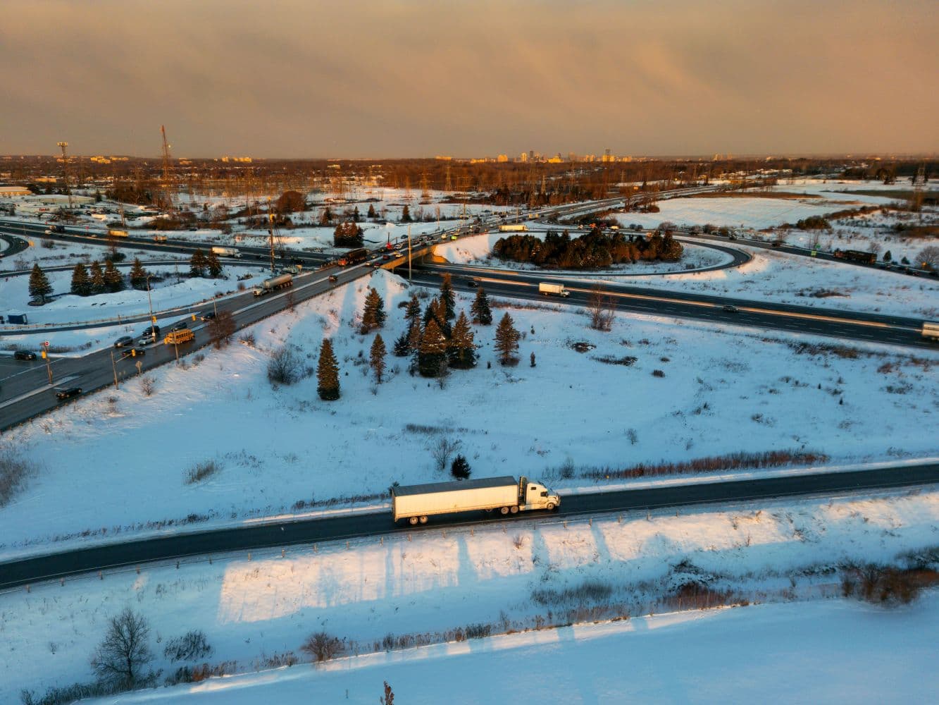 Aerial view of a semi-truck traveling along a snowy highway interchange at sunset.