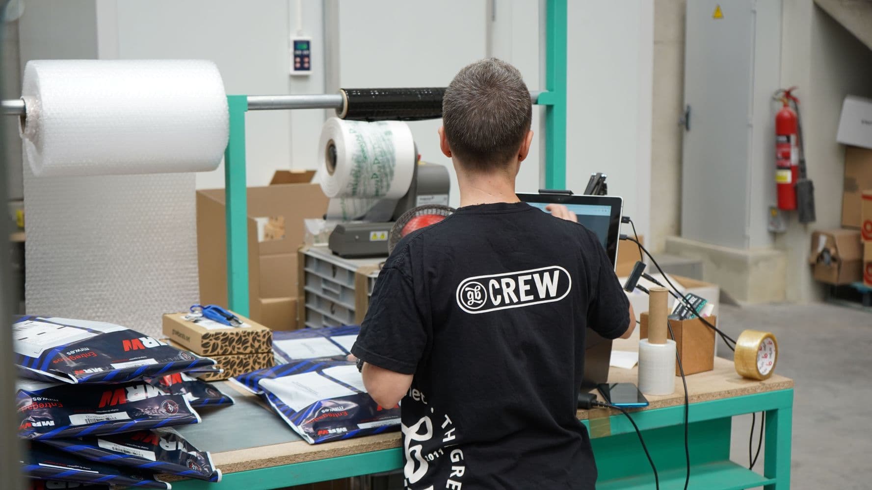 Worker processing packages at a packing station with shipping materials, boxes, and a computer.