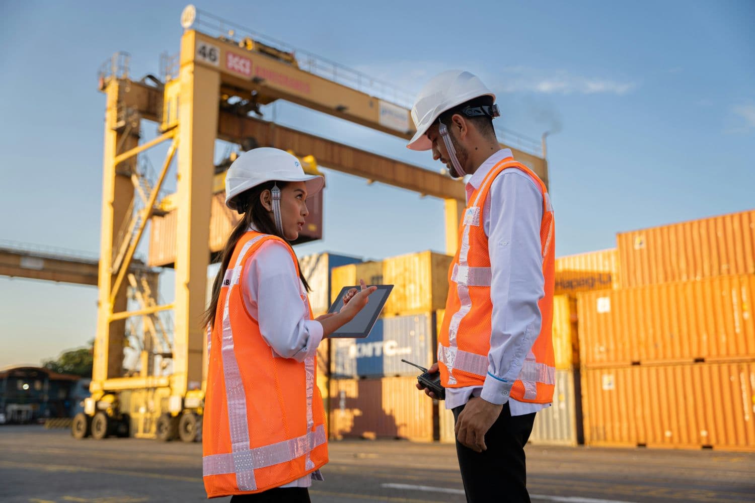Two logistics workers reviewing a tablet at a shipping container terminal.